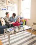 Couple sitting on a blue sofa with polka dot pillows, glass coffee table, and pink tulips in a bright living room.