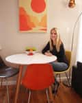 Woman sitting at a round white table with colorful chairs, a bowl of fruit, and a sunset canyon artwork on the wall.