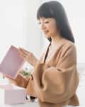 Person in a beige sweater opening a pink gift box, sitting on a light-colored sofa.