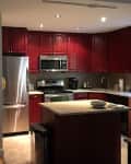 Dimly lit kitchen with red cabinets, stainless steel appliances, and a small island with a bowl and papers.