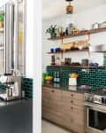 Modern kitchen with green tile backsplash, stainless steel appliances, open wooden shelves, and a coffee machine.