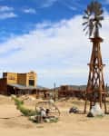 Desert landscape with a rustic wooden building, windmill, and scattered desert plants under a blue sky.