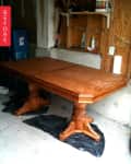 Wooden dining table with pedestal legs in a garage, surrounded by tools and storage bins.