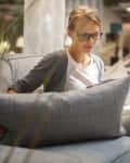 Woman sitting on a gray sofa in a store, holding a matching cushion, surrounded by home decor items.