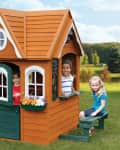 Children playing in a small orange playhouse with green doors and flower boxes in a grassy park.