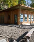 Wooden cabin with large windows by a lakeside, surrounded by trees and rocky terrain.