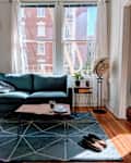Living room with a teal sofa, wooden coffee table, globe, and geometric rug, overlooking a brick building through large windows.