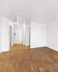 Empty room with white walls and worn wooden floor, leading to a hallway with a closed door.