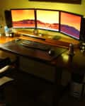 Home office setup with three monitors on a wooden desk, keyboard, mouse, lamp, and office chair.