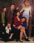 Group of nine people posing in front of a chain-link fence and brick wall, wearing casual and formal attire.