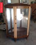 Wooden display cabinet with curved glass doors in a garage, labeled "Before" on a red tag.