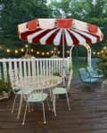 Patio with red and white umbrella, string lights, white metal table, chairs, and teal seating, surrounded by greenery.