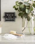 Bathroom counter with eucalyptus in a glass vase, toothbrush holder, hand brush, and "Wash Your Hands" sign.