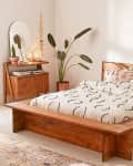 Wooden bed with patterned bedding, bedside table with mirror, plants, and decorative lamp in a cozy bedroom.
