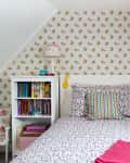Attic bedroom with floral wallpaper, single bed, colorful pillows, bookshelf, desk, and chair.