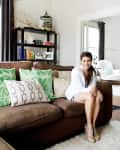 Woman sitting on a brown sofa with green and patterned pillows, next to a black bookshelf with decor items.