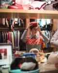 Person trying on a red hat in a vintage clothing store, surrounded by colorful clothes, shoes, and accessories.