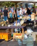 Outdoor flea market with vintage furniture, birdcages, clocks, and people browsing under umbrellas.