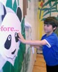 Child painting a panda mural on a wall with a roller, wearing a blue shirt.