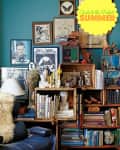 Eclectic bookshelf with books, art, sculptures, and decor against a teal wall, featuring a "Guide to the Perfect Summer" sign.
