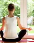 Woman practicing yoga in a bright room, sitting cross-legged on a pink mat near large windows and a potted plant.
