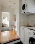 Laundry room with stacked washer and dryer, sink, and window with potted plants. Hallway leads to a sunlit room.