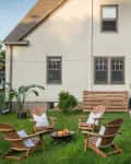 Backyard with Adirondack chairs around a fire pit, dining table under umbrella, and string lights on a house.