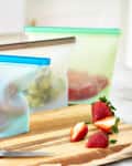 Reusable silicone bags with fruit on a wooden cutting board, knife, and bowls of sliced mango and grapes.