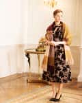 Woman in floral dress and shawl standing in elegant room with parquet floor, ornate table, and chandelier.