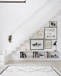 Bookshelf under staircase with framed art, books, and decor on white wall, black and white rug on wooden floor.