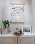 Woven wall hanging above a cabinet with potted cacti, a framed photo, and a woven tray.