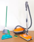 Orange vacuum cleaner with hose, green broom, teal dustpan, and sponge on wooden floor.