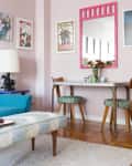 Living room with blue sofa, colorful wall art, pink mirror, and dining table with floral centerpiece.