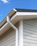 House roof corner with white siding, gutter, and downspout against a clear blue sky.