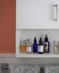 Bathroom shelf with various skincare bottles, a glass jar with cotton swabs, and floral-patterned towels hanging below.