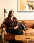 Woman sitting on a tan leather sofa with patterned pillows, next to a wooden coffee table and a floor lamp, under a vintage poster.