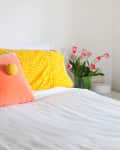 Bed with yellow polka dot pillows, blue and orange cushions, and pink tulips in a vase on a white nightstand.