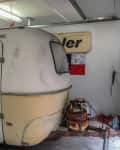 Vintage camper trailer in a garage, showing signs of wear and rust, with a propane tank attached to the front.