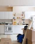 Cluttered kitchen with white cabinets, open shelves, blender, and boxes labeled "Before" in red text.