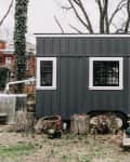 Gray tiny house with black door, white trim, and small porch surrounded by tree stumps and potted plants.
