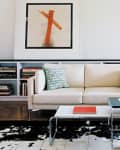 Modern living room with cream sofa, abstract wall art, cowhide rug, and stacked books on shelves.
