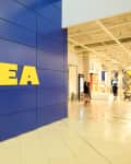 IKEA store entrance with blue wall and yellow logo, people walking inside, and visible escalator.