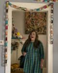 Woman in a blue plaid dress stands in a doorway with colorful paper chains, surrounded by vibrant decor and a playful poster.