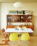 Mid-century dining room with yellow chairs, white table, wooden hutch, and pendant lamp. Decor includes vases and art.