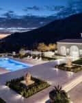 Luxury villa with illuminated pool, lounge chairs, and umbrellas, set against a mountain backdrop at sunset.