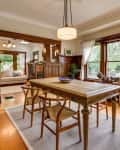 Dining room with wooden table, chairs, large windows, and view into living room with sofa and fireplace.