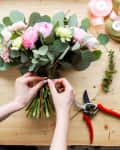 Arranging a bouquet of pink and white flowers with eucalyptus on a wooden table, surrounded by ribbons and pruning shears.