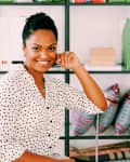 Woman in polka dot blouse sitting in front of a shelf with books, green boxes, and colorful cushions.