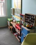Children's play kitchen with colorful appliances, toy food, and a wall poster in a carpeted room.