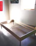 Empty wooden bed frame in a sunlit room with a small window and a floral painting on the wall.
